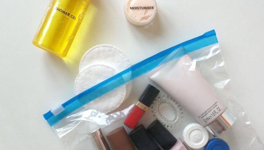 Transparenter Toppits Zipper-Beutel mit verschiedenen Kosmetikartikeln wie Creme, Lippenstift, Nagellack und Haargummi, daneben liegen Wattepads, ein gelbes Duschgel und ein kleiner Tiegel mit Feuchtigkeitscreme auf weißem Untergrund  